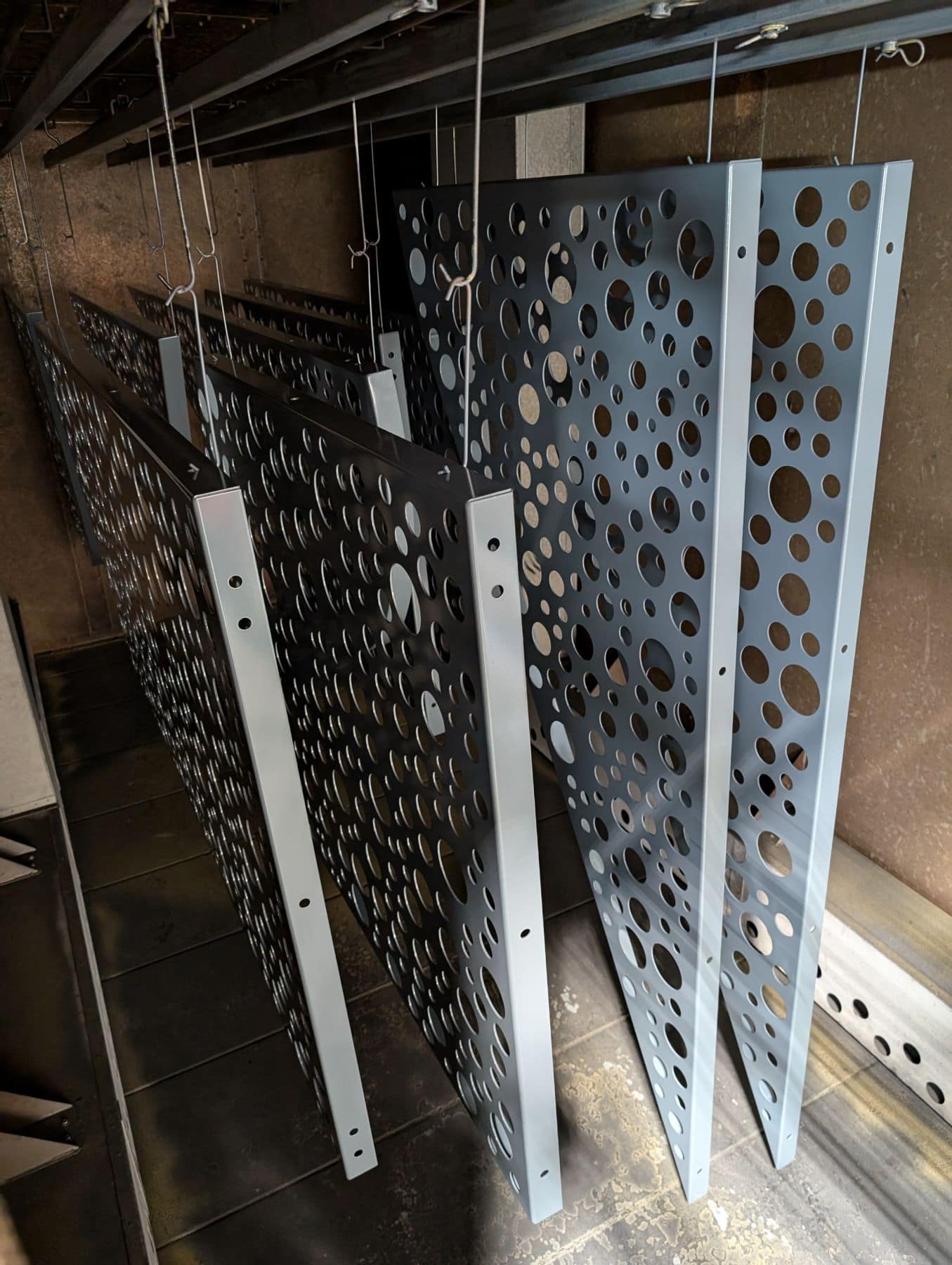 Multiple custom perforated steel panels hanging on an industrial rack undergoing the powder coating process for surface finishing.