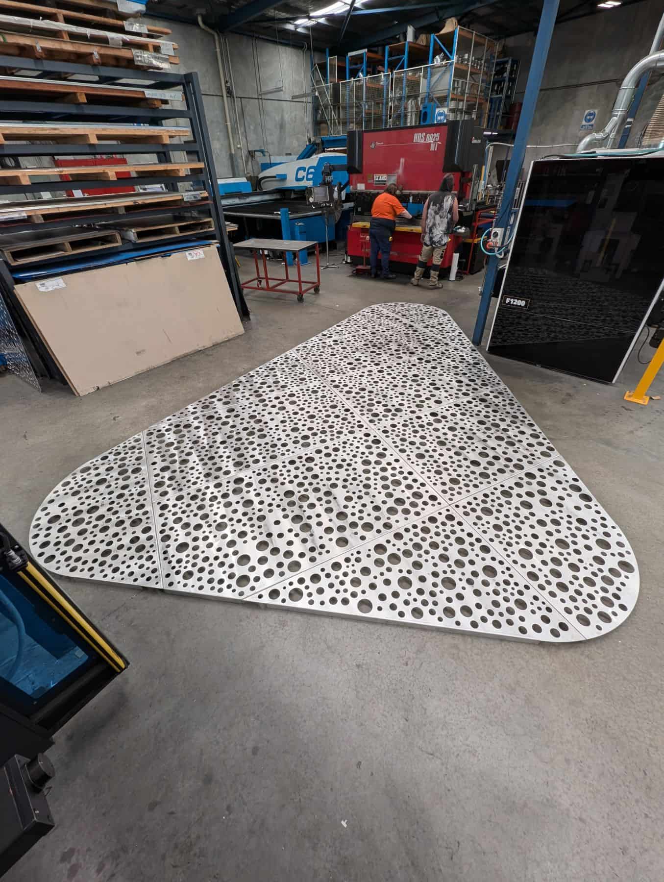 A large triangular decorative metal panel featuring a custom circular turret-punched pattern, assembled on a factory floor at Acorn Metal.